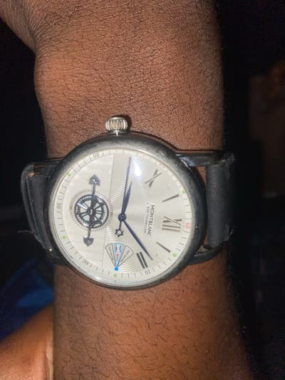 A close-up shot of a person's wrist wearing a Montblanc Exo Tourbillon watch. The watch has a silver-toned dial with Roman numerals and a sub-dial displaying a rotating mechanism with a compass-like design. The watch band is made of black leather. The person's skin is dark-skinned and has visible pores and hairs. The background is dark and out of focus, with a hint of blue and black. The location is not identifiable in the image and there is no information about the city of Wumat, Nigeria.