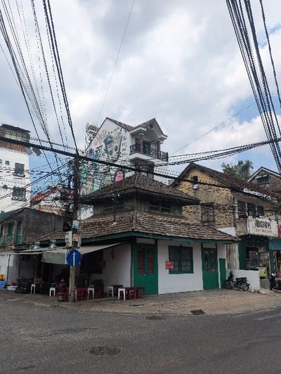 The scene depicts a street corner at 16 Lê Hồng Phong, Phường 4, Thành phố Đà Lạt, Lâm Đồng, Vietnam. A two-story building, identified as Thư Quán coffee shop, occupies the corner, featuring a white facade with dark green window frames and doors, and a multi-tiered brown tiled roof. On the ground level, several low red plastic tables with small white stools are arranged on the sidewalk under an awning.

To the left of the corner building, a weathered, unpainted two-story structure with a balcony is visible, followed by a taller white building with multiple small square windows. A utility pole stands prominently on the sidewalk, bearing a blue circular street sign with a white left-pointing arrow. A red fire hydrant is located near the pole. Various items, including blue containers, are stacked on the sidewalk next to the weathered building.

To the right, across the apparent intersection, an older, yellow-ochre building with brown windows is visible. Further right, a building with a blue facade and a partial Vietnamese sign is present. A black motorcycle is parked near the rightmost building.

The upper portion of the image is dominated by a dense, complex network of black overhead utility wires crisscrossing against a partly cloudy sky with patches of blue. The asphalt street in the foreground shows minor cracks, and a metal manhole cover is visible. No individuals are clearly identifiable or actively engaged in specific interactions; however, a faint figure is discernible in the shaded outdoor seating area to the left, and another partial figure is near the motorcycle on the far right.