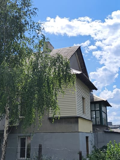 In the city of Donetsk, Ukraine, a two-story building with a grey concrete foundation and light-colored horizontal siding on the upper level is captured under a bright blue sky with scattered white clouds. A large tree with green leaves partially obscures the left side of the structure. The building features a pitched roof covered with grey corrugated material. A small, enclosed sunroom with green framing and glass panels is attached to the right side of the upper level. Windows with metal grates are visible on the lower level. The scene suggests an outdoor residential or mixed-use property.