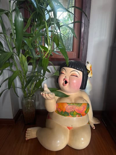 This image features a decorative statue of a plump figure, likely representing a child, in a reclining pose. The statue has dark hair adorned with a yellow and orange flower. It is wearing a colorful bikini top with a floral pattern. The figure has rosy cheeks and its eyes are closed with a wide, open-mouthed expression, giving it a joyful or surprised appearance. Its hands are outstretched, and its legs are bent. To the left of the statue, a large potted plant with long, green bamboo-like leaves is visible, with its roots submerged in a clear glass vase.
The statue is placed on a wooden surface, possibly a shelf or table, which is against a wall. A window with wooden framing is visible behind the plant, showing greenery and bright light from outside, suggesting it is daytime. The overall setting appears to be indoors, with a warm and decorative atmosphere. The statue's pose and expression contribute to a lighthearted and whimsical feel. The location context of "บ้านกาญจน์กนก 23, Thailand" suggests this might be a decorative piece in a home or establishment in Thailand. There is no visible text in the image.