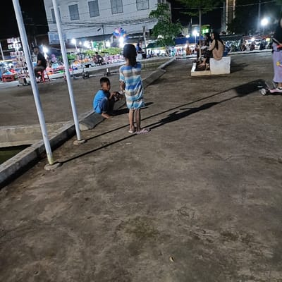 The image shows a street scene in Kaliwungu, Indonesia, at coordinates -6.962, 110.253. There are three children in the scene, one wearing blue clothing, one wearing a blue and white striped dress, and one wearing a pink and white striped shirt. The children are standing on a concrete surface, facing away from the camera. There are a few poles in the background, and shadows from the poles cast long shadows on the ground. There is also a large building in the background. There is another person sitting on a bench in the background, but they are not in focus. The scene is well-lit, suggesting that it is either daytime or nighttime with streetlights on.