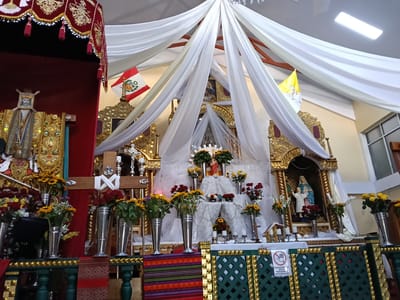 The image shows the interior of a church, likely in the city of Machupicchu, Peru. The main focus is the ornate altar, which is adorned with gold accents and religious imagery. A large, white fabric canopy drapes from the ceiling above the altar, creating a visually striking focal point. Two flags, the Peruvian flag and Vatican City flag, are displayed prominently on either side of the altar. Several silver vases filled with various flowers, including roses and sunflowers, decorate the space in front of the altar. A wooden cross is present, draped with white fabric. A sign is affixed to the front of the altar, with some text and a red circle with a line through a hand. Statues of religious figures are visible within ornate alcoves on the altar. A patterned fabric covers the space in front of the altar, and a wooden railing with gold accents borders the altar.