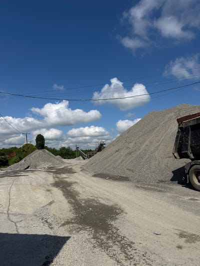 This image depicts an outdoor industrial scene, likely a gravel pit or construction material yard, under a bright blue sky with scattered white clouds. The foreground is dominated by a dirt and gravel road, with areas of darker, wet patches indicating recent water presence. To the right, a large pile of grey gravel rises steeply. A portion of a dark-colored dump truck is visible on the far right, with its tire and lower body in frame. In the middle ground, there are more piles of gravel, along with some industrial equipment, possibly conveyor belts, suggesting an active operation. A single, tall green tree stands out amongst the gravel piles. In the background, lush green trees form a treeline, and a power line with several insulators stretches across the upper portion of the image. The overall impression is one of a working industrial site.