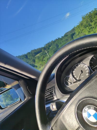 The image shows the interior of a car, likely a BMW, with a focus on the steering wheel and dashboard. The steering wheel is black leather with the BMW logo prominently displayed in the center. Part of the speedometer is visible, indicating a speed of approximately 50 km/h. To the left, a side mirror reflects a sunlit scene of trees and a road. Through the windshield, a clear blue sky with a few power lines is visible, above rolling green hills and trees. The overall impression is of driving through a rural or natural landscape on a sunny day. The image was taken from inside the car, looking forward and slightly to the right. There is no indication of the specific location in Taran, Bulgaria.
