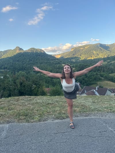 A woman is performing a yoga pose, possibly Natarajasana or Dancer's Pose, on a paved surface. She is standing on her right leg with her left leg bent and lifted behind her. Her arms are extended outwards, parallel to the ground. She is wearing a white tank top and a dark denim skirt. The background features a lush, green mountainous landscape under a partly cloudy blue sky. Several houses with red-tiled roofs are visible nestled in the valley. The image appears to have been taken in a rural or semi-rural setting outside of Saint-Claude, France.
