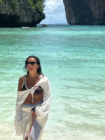 A woman stands in shallow, turquoise water near a beach with large limestone cliffs in the background. She is wearing sunglasses, a dark bikini top, a white cover-up tied at the front, and a light-colored sarong. She is holding a patterned bag in her right hand. The water is clear and calm, with some ripples on the surface. The sky is overcast with some patches of blue. The location is likely a beach in Ao Nang, Thailand, known for its limestone karst formations.