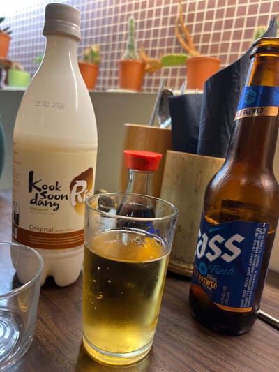 The image displays a close-up, eye-level view of a table setting featuring multiple beverages. On the left, a mostly full white plastic bottle of "Kook Soon Dang Makgeolli Original Rice Wine" with a "25.02.2026" date printed near its neck is present. The foreground also shows the rim of an empty clear glass. Centrally positioned is a clear glass containing a light amber, effervescent liquid, consistent with beer, filled approximately two-thirds. To the right, an open brown glass bottle of "Cass Fresh" beer (330ml, 4.5% alc/vol) stands next to the filled glass. Its blue label states "PRODUCT OF SOUTH KOREA" and explicitly identifies its distributor as "L'ORIENTAL BREWERY CO. LTD. 33 RUE DE LA POMPADOUR 94550 CHEVILLY LARUE FRANCE." In the background, a small clear condiment bottle with a red cap is visible, alongside two bamboo-textured cylindrical holders, one of which contains black paper napkins and silver utensils. The backdrop consists of a light brown or beige tiled wall with a grid pattern, behind which several small potted cacti and succulents in orange and green pots are arranged on a narrow shelf. The table surface is dark brown wood with visible grain. No individuals are depicted in the scene.