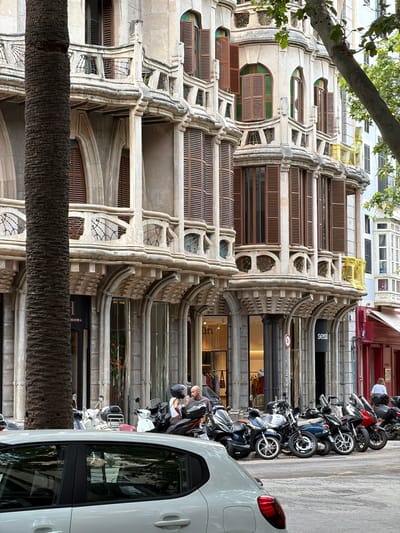 The image depicts a street scene in Palma, Spain, featuring the intricate architecture of a building with ornate balconies and shutters. In the foreground, a white car is parked on the street, reflecting the surrounding environment. Adjacent to the building, a row of motorcycles and scooters are parked along the sidewalk. Two individuals are visible near the motorcycles, one appearing to be a woman wearing a white tank top and a helmet, and the other a man in a dark shirt, both engaged in conversation. The building itself exhibits classical architectural elements, with decorative carvings and arched windows. A large palm tree trunk is prominent in the left portion of the frame, partially obscuring the building's facade. The overall atmosphere suggests a bustling urban environment on a bright day.