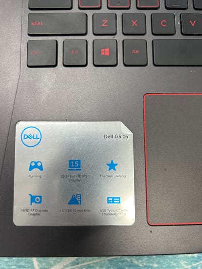 The image shows a close-up view of a Dell G5 15 laptop keyboard and a sticker with its specifications. The keyboard keys are black with red lettering and symbols. Visible keys include Shift, Ctrl, Fn, Z, X, C, V, a Windows logo, and Alt. The sticker, made of brushed metal, is placed on the palm rest. It features the Dell logo, the text "Dell G5 15", and icons with accompanying text describing features such as "Gaming", "15.6" Full HD IPS Display", "Thermal Cooling", "NVIDIA® Discrete Graphic", "< 1" / 24.95 mm thin", and "USB Type-C™ with Thunderbolt™ 3". The laptop is situated on a textured grey surface, likely a desk or table, with a thin red border visible around the touchpad area. The background is dark and out of focus. The image composition is a still life, likely taken indoors, and does not contain any information about the city of Kamianske, Ukraine.