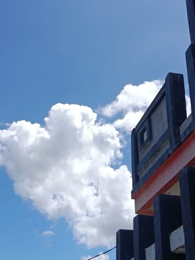 The image shows a low-angle shot of a concrete structure against a bright blue sky with fluffy white clouds. The structure is dark blue and grey with a prominent orange band. It appears to be a section of a building or a decorative element, possibly a fence or part of a rooftop. The geometric design of the structure, with its rectangular and square elements, suggests a modern or possibly brutalist architectural style. The lighting indicates it's daytime, with the sun casting shadows that define the contours of the structure. The environment is outdoors, with no other structures or natural elements visible besides the sky and clouds. The location is identified as Lebbae, Indonesia. There are no people or animals visible in the image. The image is a static, outdoor scene captured from a specific perspective.