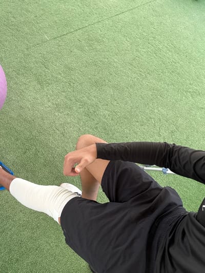 An overhead shot displays a person on a green surface, likely artificial turf. The individual wears black long sleeves, black shorts, and white athletic shoes. Their right leg is heavily wrapped with white tape or bandages from the ankle to the upper thigh. A purple exercise ball is partially visible in the bottom left corner. The person's left hand rests on their right thigh.