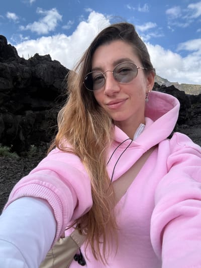 A young woman with long, wavy blonde hair, wearing round sunglasses and a pink hoodie, takes a selfie in an outdoor environment. She is smiling and looking at the camera. Behind her, dark, rocky terrain is visible, along with a cloudy blue sky. A beige strap is visible across her chest, and a white layer of clothing is visible beneath the hoodie. The scene appears to be a natural, possibly volcanic landscape. The location is identified as Kintamani, Indonesia.
