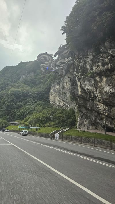 The media file shows a scenic view of a mountain landscape with a road running alongside it. The mountain is covered in lush green trees and features a large, intricate rock formation that resembles a sculpture or natural carving. The sculpture appears to depict mythical creatures or figures. 

A paved road with double white lines runs along the base of the mountain. There are a few vehicles visible on the road, including white vans and cars. A grassy embankment with a black metal fence separates the road from the mountain. Signs with text are present near the embankment, though the text is not clearly legible.

The weather appears overcast, with a grey sky. The time of day is difficult to determine precisely, but the lighting suggests it could be daytime. The overall scene evokes a sense of natural beauty and possibly a tourist attraction due to the prominent sculpture and the presence of a road and vehicles. The location is Tsementnyi, Russia.