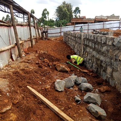 This image captures an active construction site in Borabu Chitago ward, Kenya, under an overcast sky during daytime. The foreground is dominated by reddish-brown earth scattered with numerous large, rough-hewn stones and smaller rocks.
Three individuals are visible, all engaged in work. In the immediate foreground right, a worker wearing a yellow-green high-visibility vest (with partially visible text "FIBER SOLUTI") over a light shirt and dark trousers is bent over, holding a green-handled shovel, appearing to be digging or moving earth and stones. To their right, a partially completed wall made of rough-hewn stones is visible, with mortar visible between the stones.
On the left side of the frame, a structure with a metal roof frame and corrugated metal sheet walls is being erected or reinforced. Another worker in dark clothing is bent over near this structure, working close to a wheelbarrow or similar equipment. Further in the background, a third person, wearing a patterned shirt, stands near the ongoing stone wall construction.
The background reveals a mix of lush green vegetation, including tall, slender trees and bushier plants, typical of the region. Beyond these, several residential buildings with corrugated iron roofs and brick walls are discernible. On one distant, faded building, faint text "WOMEN SELF-HELP SHOP" can be seen. The overall scene depicts ongoing manual labor and construction activities in a rural or semi-urban setting.