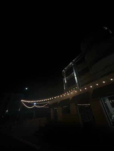 This media file depicts an outdoor scene at night, likely in Chernomorets, Bulgaria. The primary subjects are strings of small, warm-toned lights draped along an elevated structure, possibly a balcony or roofline. These lights illuminate parts of the surrounding environment, revealing architectural elements such as beams, railings, and windows. The scene is predominantly dark, indicating it is nighttime. A faint, bright circular object, possibly the moon or a distant light source, is visible in the black sky. The overall mood is serene and festive, evoked by the decorative lighting. No people are clearly discernible in the image, and no text is visible. The perspective suggests the photographer is looking upwards or from a slightly elevated position.