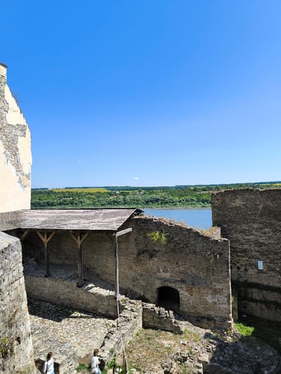 This image captures a section of the Khotyn Fortress in Khotyn, Ukraine, observed from a higher vantage point on a clear, sunny day. The scene primarily features ancient stone walls and structures, with the Dniester River visible beyond.

In the foreground and mid-ground, the fortress exhibits rugged stone architecture. To the left, a substantial, light-colored stone wall, exhibiting areas of peeled plaster and exposed masonry, forms a prominent vertical element. Centrally, a covered structure with a dark, shingled wooden roof is supported by three vertical wooden posts, providing shelter over an interior paved area. Adjacent to this and extending to the right, various stone walls and foundations show signs of wear and partial collapse. An arched, dark opening is visible at the base of a wall to the right of the wooden shelter, and a small, rectangular opening is present in a wall further right. The ground within the fortress consists of uneven stone paving interspersed with patches of dry grass and scattered debris.

Two individuals, appearing to be adult females, are partially visible in the lower-left corner of the frame. They are dressed in light-colored clothing and are positioned on the uneven ground, facing generally away from the viewer. No direct interaction between them is discernible.

Beyond the fortress walls, the blue expanse of the Dniester River flows, bordered by a distinct band of dark green trees and vegetation. In the distant background, rolling hills extend towards the horizon, displaying large patches of bright yellow agricultural fields, likely sunflowers, intermingled with areas of green foliage. The sky above is uniformly clear and blue, with no discernible clouds.