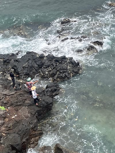 The scene depicts a rocky coastline in Desa Batu Layar Barat, Indonesia. Three individuals are engaged in fishing from dark, jagged rock formations. The person on the far left is dressed in dark clothing and stands on a rock, holding a fishing rod extended towards the water. The central figure, wearing a patterned shirt and a green and white helmet or hat, stands on an adjacent rock, holding a fishing rod and gesturing with their right arm towards the water. The person on the right is attired in a light-colored long-sleeved shirt, dark pants, and a wide-brimmed straw hat, positioned on a rock with a fishing rod cast into the ocean. The water is a muted green-blue, with white foam from waves breaking against the rocks and receding. Light-colored debris is visible floating on the water's surface near the shore.