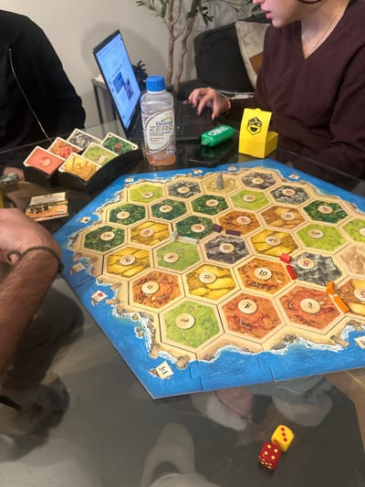 A Catan board game sits on a glass table, partially surrounded by two people. A tablet displays a webpage, next to a bottle of Electrolyte Zero drink. Resource cards are visible near a person's hand, close to a Catan player aid card. Dice lie off the edge of the board. The board features resource hexes with number tokens. A yellow resource card box, and two green ENEO containers sit nearby.