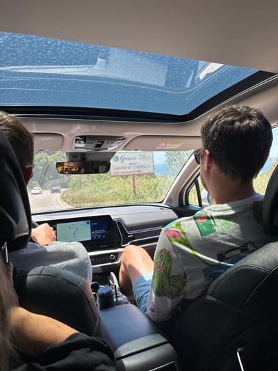 The interior of a vehicle traveling on a road in Kunje, Montenegro, is shown. Two adult male passengers are visible: one in the front passenger seat and one in the driver's seat. The passenger is leaning forward looking out of the windshield. The GPS is displaying a map with a blue line indicating the route. The rear view mirror is showing the road and a sign. The windshield has streaks of water on it.