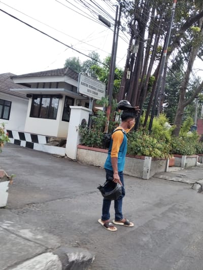 This media file shows a person walking on a paved road in front of a building. The person is wearing a blue and orange shirt, dark blue jeans, and sandals. They are carrying two helmets, one on their head and one in their hand. A backpack is slung over their shoulder. The building appears to be an office or government facility, with a sign that reads "KANTOR BERSAMA SAMSAT KOTA CIMAHI" in Indonesian, indicating it is a joint Samsat office in Cimahi City. The sign also mentions "DIREKTORAT LALU LINTAS POLDA JABAR" (Traffic Directorate of West Java Regional Police) and "DINAS PENDAPATAN PROPINSI JAWA BARAT" (West Java Provincial Revenue Agency). The scene suggests a daytime setting, possibly late morning or early afternoon, with overcast or bright conditions judging by the lighting. The person appears to be walking away from the viewer, with their back slightly turned. The surroundings include trees, electrical poles with numerous wires, and some landscaping in planters along the building's facade. The road surface is asphalt with some minor cracks. The overall impression is a casual street scene in an urban Indonesian environment.
