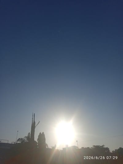 The image depicts an outdoor scene at sunrise. The sun is prominently featured, creating a bright glare and lens flare effects across the sky. Silhouetted against the bright sky are trees and a tall, thin structure that resembles a communication tower. In the foreground, a wall with barbed wire and some architectural elements are visible, partially obscured by the light. The sky is a clear, deep blue, transitioning to a lighter hue near the horizon. The overall impression is one of early morning light. The date and time stamp "2026/6/26 07:29" is visible in the bottom right corner, indicating the image was taken in Embakasi, Kenya.