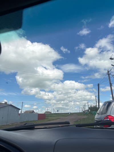 This is a photograph taken from the interior of a vehicle, looking out through the windshield. The foreground shows the dashboard and windshield wipers. The scene outside is a rural or semi-rural landscape under a bright blue sky with fluffy white clouds. In the middle ground, there is a dirt road leading into the distance. To the left, there are several buildings, including a gray corrugated metal garage or shed and a red and gray fence. Further down the road, there are more houses and utility poles with overhead wires. To the right, a silver hatchback car is parked or stopped on the side of the road. The location appears to be Северная, Russia, based on the text provided with the image. The overall impression is a sunny day in a village or small town setting.