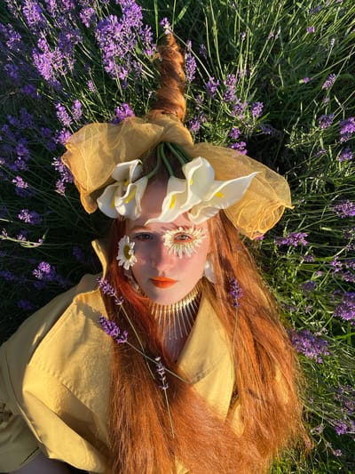 A woman with auburn hair lies in a field of lavender. She is wearing a yellow jacket and a headdress adorned with calla lilies and a daisy. Her makeup is dramatic, with orange lipstick and decorated eyelashes. The scene is illuminated by the warm glow of sunlight. The location is described as being in Verkhneye Uchdere, Russia, suggesting a natural, outdoor setting.