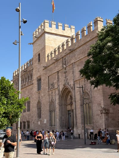 This media file captures an outdoor scene with a large, ornate stone building as the backdrop, likely a historical or religious structure due to its intricate architectural details, including arched windows and decorative carvings. The building's facade is made of light-colored stone, and its upper edges are adorned with battlements. A large green tree with dense foliage partially obscures the top left portion of the building.
In the foreground, a group of people are gathered, suggesting a public space or tourist attraction. Some individuals are standing, while others appear to be seated. A flag with red and yellow stripes is visible on a flagpole to the left of the building. There's also a modern streetlamp with multiple lights, indicating an urban environment.
A group of musicians, possibly a string quartet, are performing in front of the building. One person is playing a cello, and others are holding violins. They are dressed in casual attire. A small crowd of onlookers has gathered to watch the performance. The people in the crowd are dressed in summer clothing, suggesting warm weather. The lighting indicates it is daytime, with sunlight casting shadows on the ground and the building.
In the bottom right of the frame, a man is standing and looking at something he is holding, possibly a map or a brochure. He is wearing a black polo shirt and light-colored shorts. The overall atmosphere seems relaxed and pleasant, with people enjoying the music and the surroundings. The location is identified as Alboraia / Alboraya, Spain. No visible text is discernible in the image.