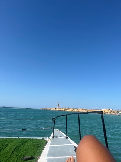 The image depicts a view from a boat or dock, likely on the water off the coast of Unknown, Venezuela. The sky is clear and blue, occupying a significant portion of the frame. A narrow strip of land runs along the horizon, where a lighthouse can be identified. The water is greenish-blue and textured with small waves. A black metal railing and the edge of a deck are visible on the right side of the image. The deck has patches of artificial grass. A person's bare foot and part of a leg are present in the upper right corner.