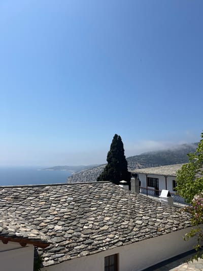 The image shows a panoramic view from a rooftop in Moni Archaggelou, Greece (latitude: 40.597, longitude: 24.701). The scene features a stone-tiled roof, white-washed walls, and a small evergreen tree, all positioned on a cliff overlooking a vast blue sea. The background shows a hazy coastline and a sky with a light blue hue. There are no people in the image. The environment suggests a serene and idyllic location.