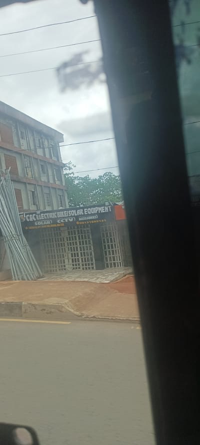 The image, taken from within a vehicle, shows the exterior of a business named "C&C Electric Bike/Solar Equipment". The business provides services related to solar power, CCTV, and bike accessories, and is located at 5 Mission Hill, opposite Okigwe Park, in Umuahia, Abia State, Nigeria. The phone number for the business is 09131665131.
The building appears to be a multi-story structure with what seem to be closed window coverings. A pile of metal poles or pipes rests on the left side of the building. The business has metal security bars in front of the storefront. The image is somewhat obscured by a dark object in the foreground, most likely part of the vehicle interior. The sky appears overcast. A road runs in front of the building.