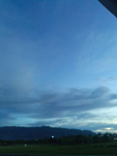 A blurry, outdoor, low-angle full shot captures a twilight sky over a dark mountain range and fields in Meunasahmanyet, Indonesia. The sky is a gradient of deep blue to lighter blue. A dark, irregular line across the top of the image suggests an overhang or structure. The middle ground shows a dark, out-of-focus mountain range with a faint, circular light source to the right of center. Below the mountains, out-of-focus green fields and dark trees are visible. Another faint, circular light source is in the field to the left of the mountain range.