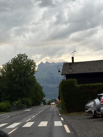 The image depicts a road scene in Saint-Gervais-les-Bains, France, likely in the late afternoon or early evening given the overcast sky and ambient light. A paved road with white dashed lines and a pedestrian crossing is visible in the foreground, leading towards a distant mountain range. On the right side of the road, there is a residential building with a dark tiled roof, a chimney with an antenna, and a tall, manicured hedge. Parked on the shoulder next to the hedge are two silver SUVs, one of which is partially obscured. On the left side of the road, a large tree with dense green foliage dominates the mid-ground, and beyond it, a darker wooded area and more distant mountains can be seen. Two cars with their headlights on are driving away from the viewer down the road. The sky is filled with heavy, grey clouds.