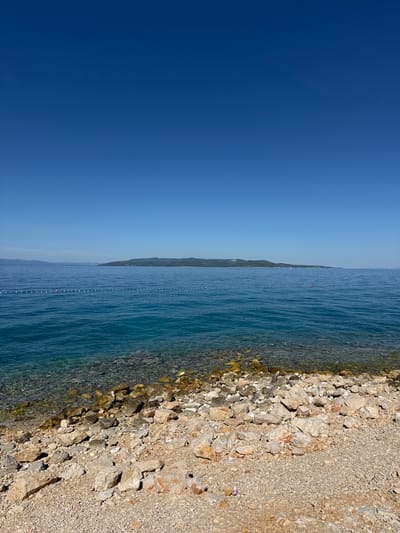 The image shows a clear, blue sky above a calm, turquoise sea. In the foreground, a rocky beach with small pebbles and larger stones lines the shore. The water is shallow near the beach, revealing the rocky seabed. A white rope with buoys separates the swimming area from the open sea. In the distance, a long, hilly island is visible under the clear sky. The scene is one of a serene coastal landscape, likely in a Mediterranean setting. Based on the visual cues such as the island formation and the characteristic Mediterranean vegetation visible on the distant shore, this scene is highly consistent with the coastline of Makarska, Croatia, a city known for its beautiful beaches and proximity to islands.