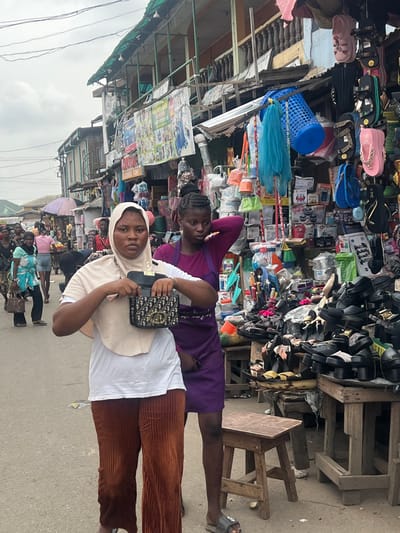 The image depicts a bustling outdoor market street in Egan, Nigeria, under an overcast sky. In the foreground, a person wearing a light beige hijab over a white t-shirt and brown corduroy pants holds open a small, dark handbag featuring a gold-colored "CD" style logo, looking directly at the camera with a neutral expression. To their right and slightly behind, another individual with braided hair, wearing a solid purple dress or apron-style garment, stands near a market stall, their left hand touching the back of their head. They wear a single dark slide sandal on their visible foot.
The background is densely packed with market stalls and activity. To the immediate right, a wooden stall displays numerous shoes, sandals, and flip-flops, predominantly black, stacked and arranged on trays and wooden structures. Above this, household items such as plastic buckets, brushes, and containers are displayed, some hanging, alongside brightly colored flip-flops and a blue plastic laundry basket. Further into the background, multiple multi-story buildings line the street, featuring weathered facades, exposed wooden balconies, and corrugated metal awnings. Various signs, including one partially visible with "BLESSED TOWN," are affixed to these structures. Numerous other individuals are visible throughout the street, walking and engaging in market activities. The street pavement is a light gray. Overhead, utility lines crisscross the sky. Two small wooden stools rest on the pavement in the foreground.