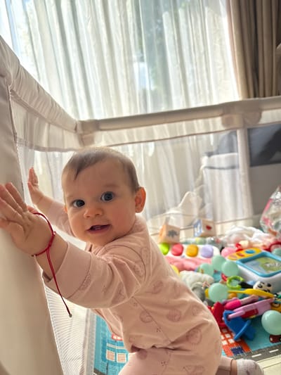 A baby wearing pink pajamas is leaning over the edge of a crib. The baby is looking at the camera and smiling. The crib is white with a mesh side. Toys are visible inside the crib. The location is Veghel, Netherlands. Curtains are visible in the background.