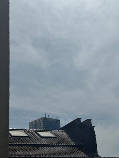 This media file features an upward-looking view of a cloudy sky with two buildings partially visible on the right. The building in the foreground is a dark-tiled structure with two square windows. The roofline of this building is prominent and features a stepped, angled design. Behind it, a taller, glass-facade building with a rectangular shape is visible. This second building appears to have numerous antennas or communication equipment on its roof. The sky is overcast, suggesting a cloudy day, and the lighting is diffused. The overall impression is a view from a lower vantage point looking up towards the sky and surrounding urban architecture. There is no visible text in the image. The setting appears to be an urban environment, consistent with the provided location context of Molenbeek-Saint-Jean - Sint-Jans-Molenbeek, Belgium. The image captures a slice of the city's skyline, focusing on the interaction between architectural forms and the atmospheric conditions.