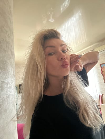 A young woman with blonde hair pulled back from her face is posing for a selfie. She is wearing a black shirt and has her left hand touching her cheek, with her lips puckered. The background appears to be an indoor setting, possibly a home or hotel room, with a textured wall on the left and a window with blinds on the right. There are reflections on the ceiling, suggesting it is a smooth, possibly glossy surface. The overall lighting is soft and diffused. The image does not contain any specific geographical markers that would indicate the location is Armavir, Russia.