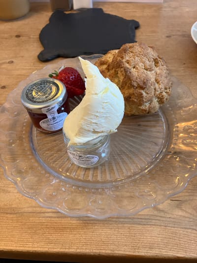 A clear, scalloped-edge glass plate containing a cream tea service rests on a light-colored wooden table in Ambleside, United Kingdom. The plate displays one large, golden-brown scone with a textured crust. To the scone's left, a small glass jar holds a substantial mound of off-white clotted cream. Adjacent to the cream, another small glass jar contains red strawberry jam, identifiable by its "WILKIN & SONS LTD STRAWBERRY PRESERVE TIPPERY ESSEX" label. A single, whole, ripe red strawberry is positioned next to the jam jar. In the background, a dark, irregularly shaped object, likely a coaster, is visible on the wooden surface, alongside blurred lighter elements consistent with a cafe interior.