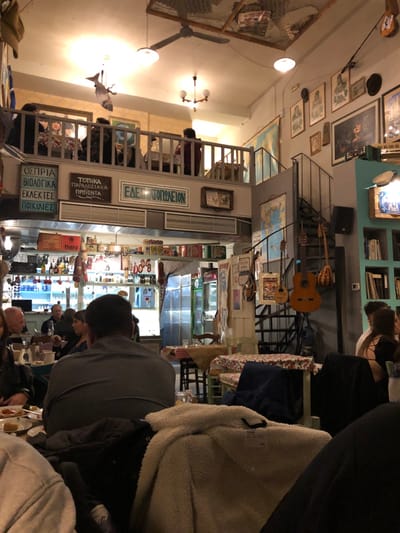 The image captures the interior of a bustling, rustic restaurant or taverna, likely in the evening given the artificial lighting. The scene is viewed from a low perspective, as if from a diner's seat, looking across and into the establishment.
Numerous patrons are scattered throughout the space. In the foreground, the back of a person wearing a grey shirt is visible, seated at a table with others whose faces are partially obscured. A cream-colored, fleece-lined jacket is draped over a chair or person in the immediate foreground. The restaurant features a multi-level layout, with a mezzanine or upper dining area visible on the left side of the frame, accessible by a staircase. More diners are seated at tables in this upper section.
The establishment is richly decorated with an eclectic mix of items, creating a cozy and lived-in atmosphere. Walls are adorned with various framed pictures and artworks, including what appear to be landscapes and portraits. Musical instruments, specifically guitars and a bouzouki or mandolin, hang decoratively near the staircase. A large world map is displayed on a wall near the mezzanine. Overhead, a wooden ceiling fan and several light fixtures, including chandeliers and simple hanging bulbs, illuminate the space. A decorative fish ornament also hangs from the ceiling.
In the mid-ground, behind the ground-floor diners, there's a counter or shelving unit displaying various products, including bottles and packages. Visible text on some of the signs below the mezzanine railing is in Greek: "ΕΛΕΥΘΕΡΟΠΩΛΕΙΟΝ" (Eleftheropoleion), and listings such as "ΤΟΠΙΚΑ ΠΑΡΑΔΟΣΙΑΚΑ ΠΡΟΪΟΝΤΑ" (Local Traditional Products), "ΟΣΠΡΙΑ ΒΙΟΛΟΓΙΚΑ" (Organic Legumes), and "ΕΚΛΕΚΤΕΣ ΠΟΙΚΙΛΙΕΣ" (Selected Varieties). Other brand names like "Pampers" and "Haig Club" are discernible on packages and bottles on the shelves. The overall impression is one of a warm, inviting Greek eatery where people are enjoying meals and conversation.