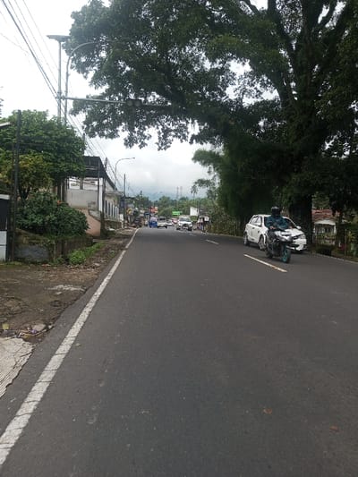 The image presents a forward-facing perspective of a paved two-lane road in Ariyalai East, Sri Lanka, under an overcast sky. The road features a continuous white line delineating its left edge. To the left of the road, a dirt and concrete shoulder abuts a series of structures, including a weathered building and a utility pole supporting a solar-powered street lamp. Dense green vegetation, comprising shrubs and a large tree with branches extending over the road, lines both sides of the thoroughfare. In the midground, a person wearing a dark helmet and jacket operates a dark motorcycle with light blue wheel accents, positioned on the right side of the road and traveling away from the viewer. Immediately behind the motorcycle is a white four-door sedan, also moving away. In the distant background, several vehicles, including a light blue pickup truck, proceed in the same direction, with a horizontally mounted traffic signal visible above them, partially obscured by tree foliage. Mountains or hills are faintly visible on the horizon.