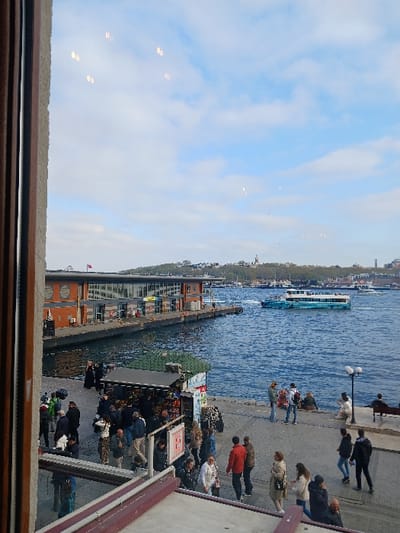 A view from a window overlooking the Golden Horn in Istanbul, Türkiye (41.022, 28.976). The image shows a ferry in the water and a number of people walking on the promenade in front of a building with a red roof and large windows. The sky is blue with a few white clouds. The image is taken from a building, likely a cafe, where the viewer can see people sitting at tables and the view through the window. Several people are looking out towards the water, which is calm and reflecting the blue sky. A few small boats are docked in the water behind the ferry. The environment appears to be a busy, bustling urban area, with the promenade, ferry, and cafe suggesting a popular tourist destination. The building in the background is likely a ferry terminal, with a red flag flying from its roof.