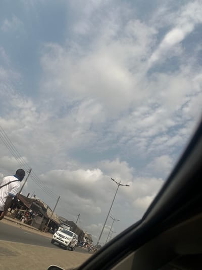 The image depicts an urban street scene in Igwuruta, Nigeria, under a partly cloudy sky. The perspective is from inside a vehicle, indicated by a dark diagonal frame element in the lower right. The upper two-thirds of the frame feature a bright sky with scattered white and light gray cumulus clouds against a pale blue background.
In the lower-left foreground, an adult male, of African descent, is seen from behind, walking away from the camera on what appears to be a roadside shoulder or sidewalk. He is wearing a white short-sleeved collared shirt with blue accents and has a dark strap, possibly from a bag, across his left shoulder.
Along the left side of the paved road, multiple utility poles with overhead power lines are visible. Behind these poles, a continuous line of single-story commercial buildings or stalls with corrugated metal roofs and various signs, including one partially visible "OSI" text, extend into the distance, indicating a market or commercial strip.
On the paved road itself, a white four-door pickup truck is observed driving towards the camera in the left lane. Further down the road, additional vehicles and pedestrians are discernible, suggesting moderate traffic activity. Parallel to the road on the right side, several tall, dark streetlights with dual lamp heads are positioned, extending into the background. The overall scene suggests a busy daytime environment along a main road.