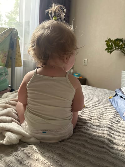 A toddler with blonde hair tied into a high ponytail sits on a bed facing away from the camera. The toddler is wearing a white tank top and a diaper. The bed is covered with a textured grey blanket and a cream-colored fluffy blanket is draped over the left side. In the background, a window with sheer curtains allows natural light to enter the room. To the left of the window, a baby crib with a patterned quilt is visible. A light switch is mounted on the wall behind the toddler. A potted plant with green leaves sits on a nightstand to the right. The overall setting suggests a domestic environment, likely a bedroom, in the city of Sayanogorsk, Russia.
