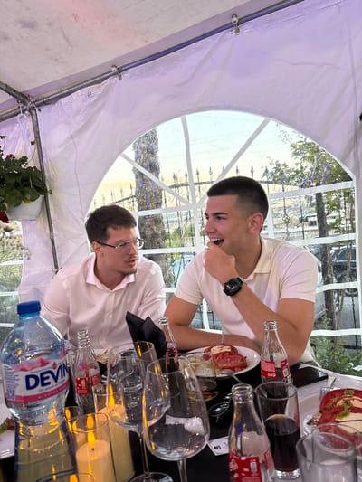 The image depicts two young men seated at a table outdoors, likely at a restaurant or event. The scene appears to be in Razgrad, Bulgaria, judging by the text on the water bottle. They are seated under a white tent with a clear arched window or opening, allowing a view of trees and foliage outside.

The man on the left is wearing a light-colored button-down shirt and glasses. He is looking to his right, seemingly engaged in conversation. The man on the right is wearing a white polo shirt and is laughing, with his hand near his mouth. He is also looking to his left.

On the table, there are several items, including a large bottle of "DEVIN" mineral water, glasses (wine glasses and tumblers), bottles of what appears to be Coca-Cola, and plates of food, one of which contains sliced tomatoes and cheese, resembling a caprese salad. There are also lit candles on the table, providing ambient lighting. The overall atmosphere suggests a social gathering or meal.