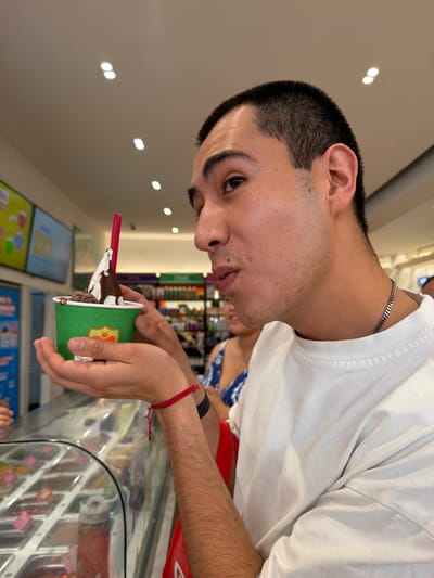 An eye-level indoor shot captures a young man in a frozen yogurt or ice cream shop in San Francisco Ocotlán, Mexico. The subject, a male with short dark hair, is seen from the chest up, looking downwards at a green paper cup he holds with both hands. He is wearing a white t-shirt, a thin silver chain, and a red string bracelet on his right wrist. His lips are slightly pursed as he gazes at the dessert.

The green cup contains a white soft-serve substance topped with dark chocolate sauce and other dark, granular toppings, possibly cookie crumbles. A red spoon is inserted into the dessert. The cup features a partial orange or yellow logo.

The background shows a modern retail environment. To the lower right, a clear glass display case with various colorful toppings is visible. To the left, two rectangular screens display bright, colorful graphics, likely advertisements for the shop's offerings, though no specific text is legible. The ceiling is light-colored with multiple recessed lights, indicating a well-lit interior, suggesting it could be daytime or evening. A blurred figure in the background appears to be wearing a blue and white patterned top and red clothing. The overall scene conveys a casual atmosphere of enjoying a sweet treat.