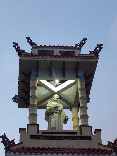 An upward-angled photograph captures the illuminated upper section of a multi-tiered bell tower, set against a light blue-grey sky. At the core of the structure, an elevated, light-colored statue of a bearded male figure, identifiable as Saint Francis of Assisi, stands within an open pavilion. The statue wears a monastic robe, holds an object resembling a book or scroll against its left chest, and has a small bird, possibly a dove, perched on its right shoulder.

The pavilion is supported by four segmented pillars, designed to mimic bamboo stalks, and features a light-colored roof structure with prominent, ornate dark-colored upturned corner decorations. A single bright, circular light fixture is mounted on the underside of the pavilion's roof, directly illuminating the statue. Below this pavilion, a section of a lower tier is visible, characterized by a roof covered in red-brown interlocking tiles and adorned with similar decorative elements at its corners. The walls of this lower tier are light-colored, featuring a dark, horizontally banded decorative frieze. This bell tower is part of the Franciscan Monastery (Tu viện Dòng Phanxicô) in Thành phố Đà Lạt, Vietnam.