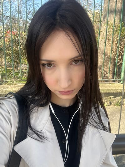 A young female is centrally positioned in a close-up, chest-up outdoor portrait. She has long, dark brown hair with a central part, brown eyes with visible eyeliner, and a fair complexion with evident blush on her cheeks. Her lips are slightly parted, and her gaze is directed straight ahead. She wears a light beige trench coat over a black button-down garment. White wired earphones are draped around her neck. A black strap, consistent with a bag, is visible over her left shoulder. The background is out of focus, revealing a green metal fence with vertical bars. Behind the fence, deciduous trees with sparse branches are visible against a clear blue sky, indicating late winter or early spring. Small red blossoms or foliage are present on the left side of the background. The scene is located in an outdoor public area within Cachan, France; no specific street or landmark is discernible.