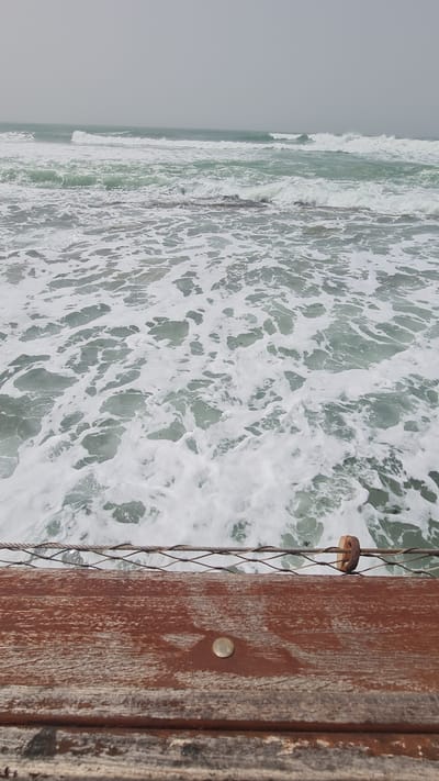 The image depicts an ocean view from what appears to be a pier or boardwalk in Nahariyya, Israel. The foreground contains a wooden structure with a metal screen or netting attached. The ocean's surface is foamy and turbulent. Waves are breaking in the distance. The sky is overcast and gray.