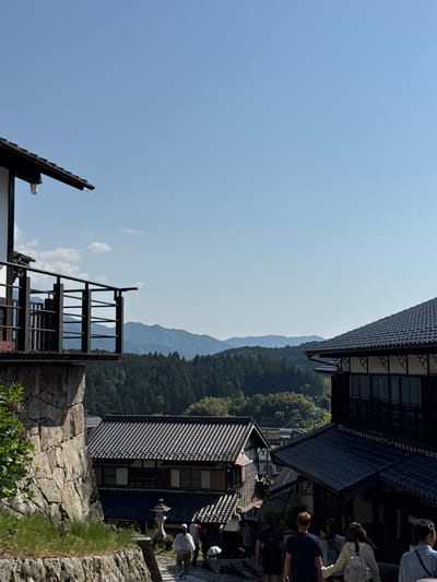 The image depicts a partial view of a street scene in the city of Nakatsugawa, Japan, characterized by traditional Japanese architecture.

Several buildings are visible, featuring dark tiled roofs and wooden frameworks. A stone wall is in the foreground and structures include a set of metal stairs, roofs, and architectural elements.

In the background, forested mountains are visible.

Several people are present in the upper right portion of the image.
